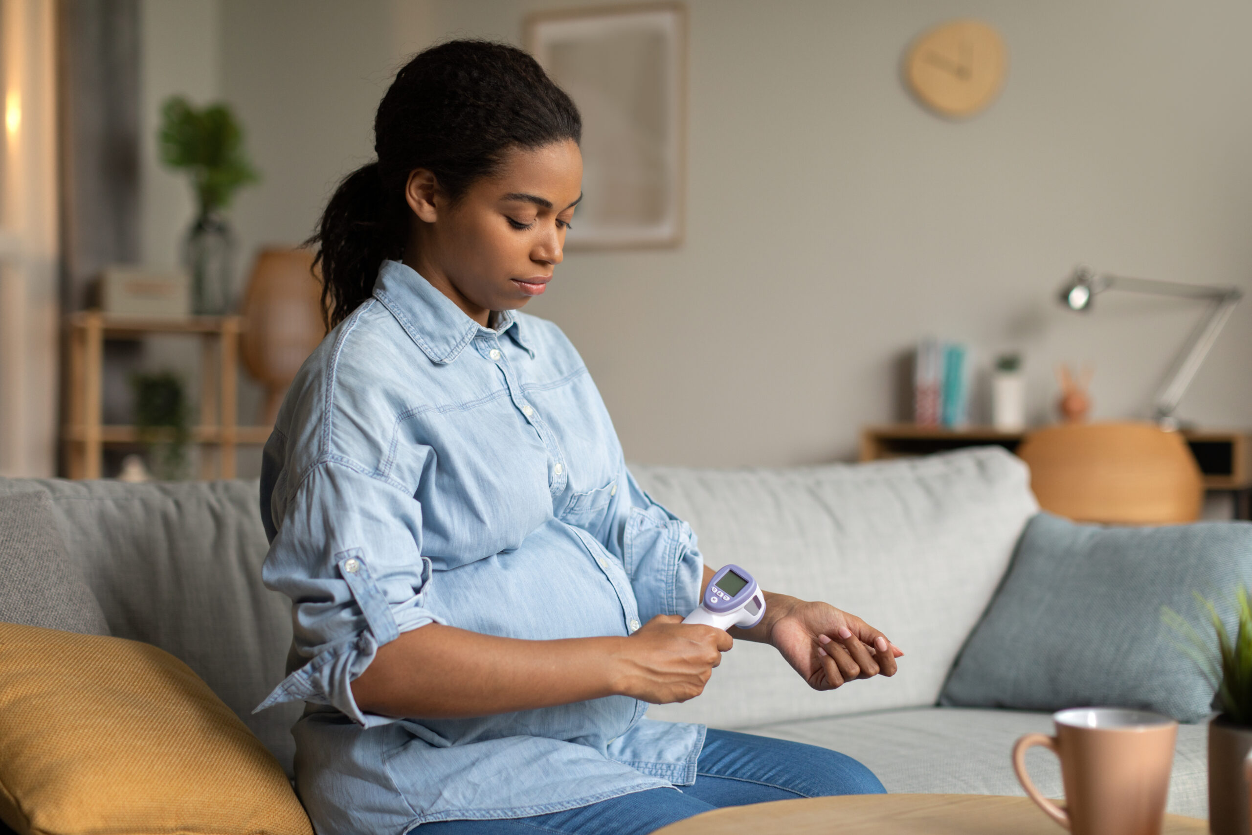 Sick Pregnant Black Woman Measuring Temperature With Non Contact Thermometer Having Fever Sitting On Couch At Home. Disease Symptom, Sickness In Pregnancy Concept. Side View