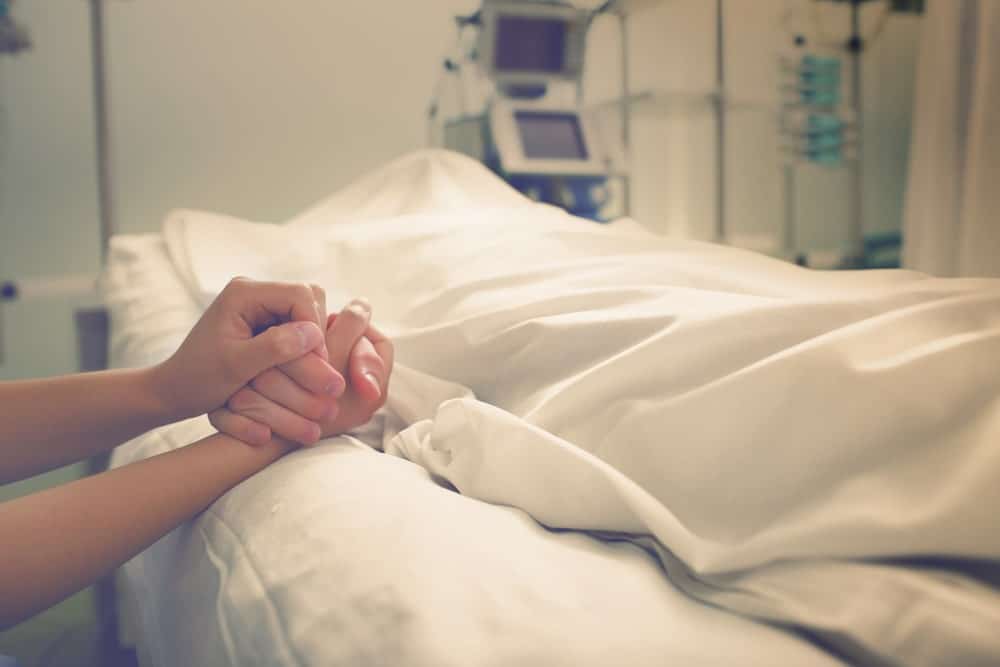 A Person Lying On A Hospital Bed And A Visitor Holding His Hand