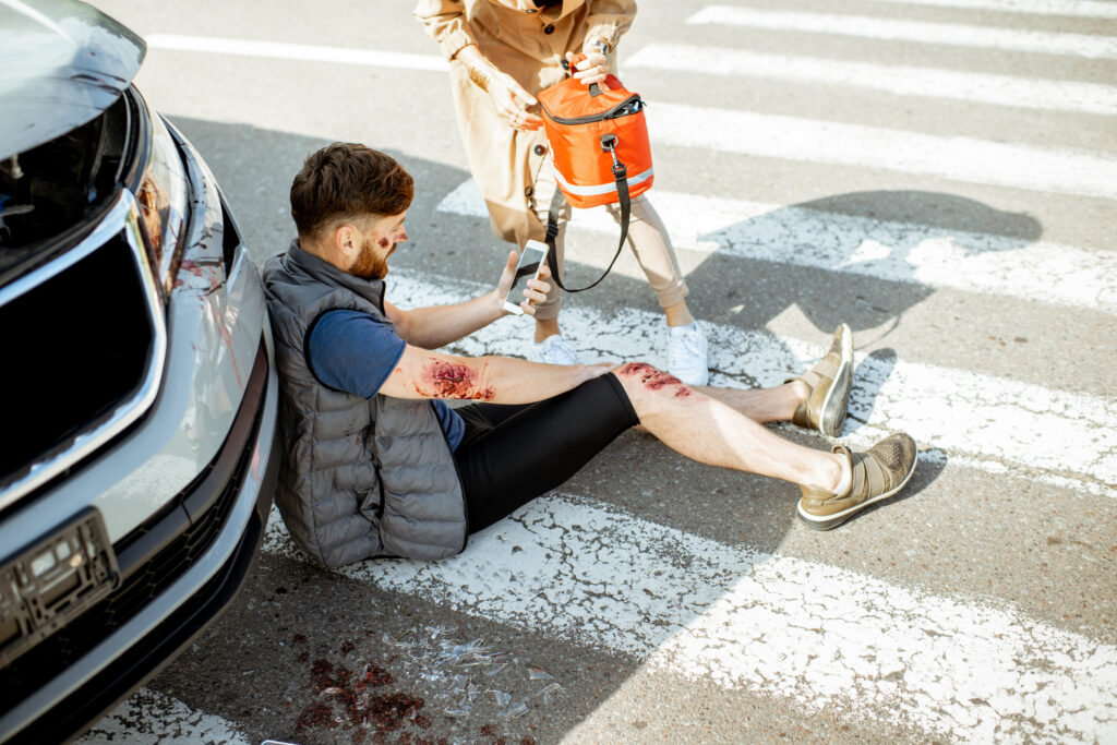 Driver hurrying with first aid kit to help injured man with bleeding wounds sitting near the car after the road accident